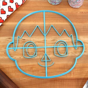 May include: A blue plastic cookie cutter shaped like a cartoon character with a smiling face, spiky hair, and large eyes. The cutter is on a wooden surface.