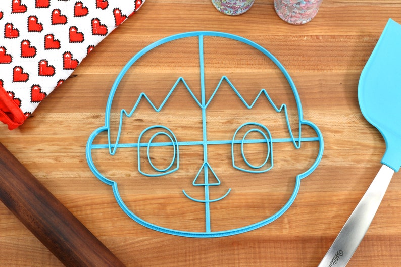 May include: A blue plastic cookie cutter shaped like a cartoon character with a smiling face, spiky hair, and large eyes. The cutter is on a wooden surface.