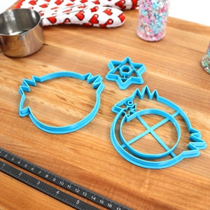 May include: Three blue plastic cookie cutters on a wooden surface. The cutters are shaped like a star of David, a round object with a cross inside, and a round object with a wavy edge.