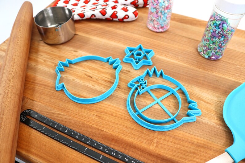 May include: Three blue plastic cookie cutters on a wooden surface. The cutters are shaped like a star of David, a round object with a cross inside, and a round object with a wavy edge.