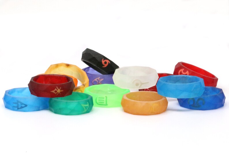 May include: A collection of 12 colorful silicone rings with different symbols and designs. The rings are stacked and arranged in a semi-circle on a white surface.