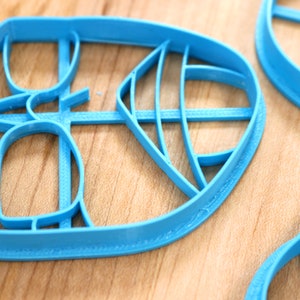 May include: Three blue plastic cookie cutters shaped like leaves. The cutters have intricate designs and are on a wooden surface.