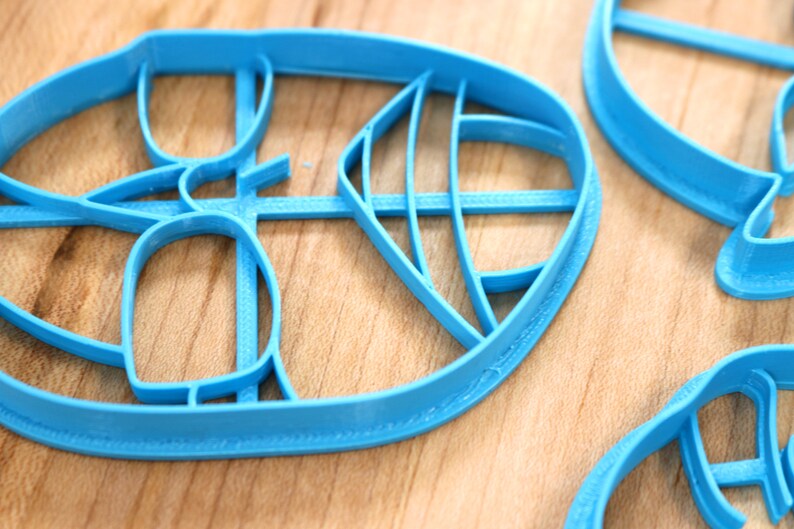May include: Three blue plastic cookie cutters shaped like leaves. The cutters have intricate designs and are on a wooden surface.