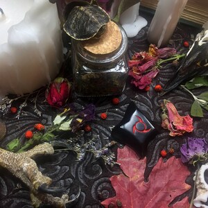 K&ouml;nnte beinhalten: Ein Stillleben mit einer gro&szlig;en wei&szlig;en Kerze, einem Glas mit Korken, getrockneten Blumen, einem Vogelsch&auml;del und einem schwarzen dekorativen Objekt mit rotem Design. Die Szene ist auf einer dunklen, gemusterten Oberfl&auml;che arrangiert.