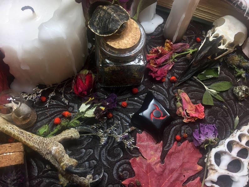 K&ouml;nnte beinhalten: Ein Stillleben mit einer gro&szlig;en wei&szlig;en Kerze, einem Glas mit Korken, getrockneten Blumen, einem Vogelsch&auml;del und einem schwarzen dekorativen Objekt mit rotem Design. Die Szene ist auf einer dunklen, gemusterten Oberfl&auml;che arrangiert.
