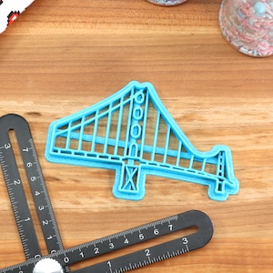 May include: A blue plastic cookie cutter shaped like the Golden Gate Bridge. The cutter is on a wooden surface.