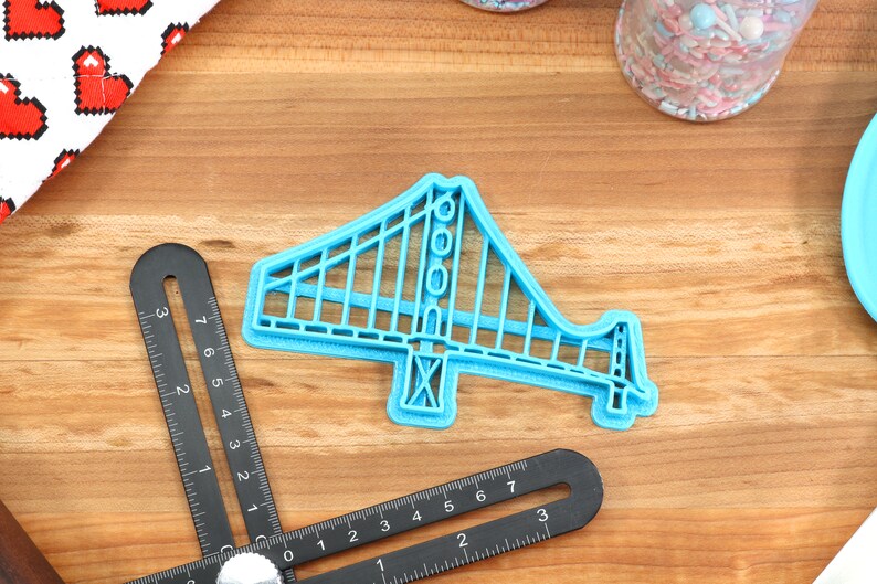 May include: A blue plastic cookie cutter shaped like the Golden Gate Bridge. The cutter is on a wooden surface.