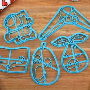 May include: A collection of five light blue plastic cookie cutters in various shapes, including a baby carriage, a onesie, a pear, a bow, and a baby bottle. The cutters are arranged on a wooden surface.