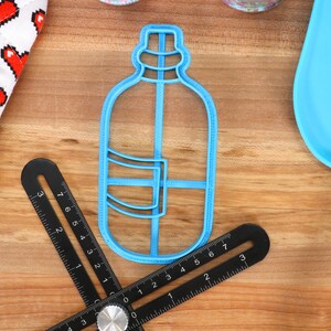 以下が含まれることがあります： 青いプラスチック製のクッキー型で、水筒の形をしています。クッキー型は、黒い定規と青いシリコン製のへらのある木製の表面の上に置かれています。