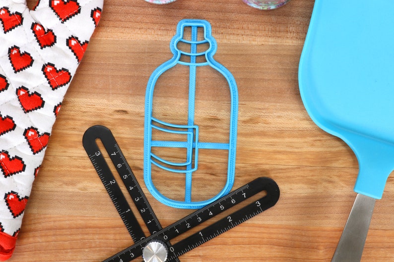 以下が含まれることがあります： 青いプラスチック製のクッキー型で、水筒の形をしています。クッキー型は、黒い定規と青いシリコン製のへらのある木製の表面の上に置かれています。