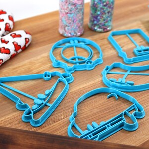 May include: A set of five blue plastic cookie cutters shaped like a car, a birthday cake, a hat, a hot dog, and a donut. The cutters are on a wooden surface.