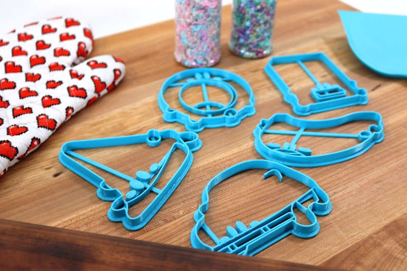 May include: A set of five blue plastic cookie cutters shaped like a car, a birthday cake, a hat, a hot dog, and a donut. The cutters are on a wooden surface.