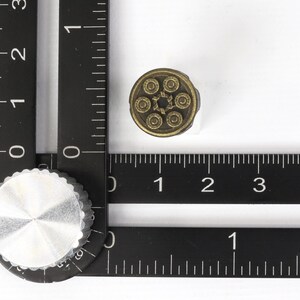 以下が含まれることがあります： 中央の星を中心に6つの円がデザインされた、小さくて丸いアンティーク真鍮色のノブ。ノブは、メートル法による測定値を持つ黒い定規の隣の白い表面に配置されています。