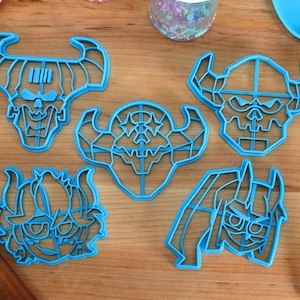 May include: Five blue plastic cookie cutters shaped like cartoon demon heads with horns. The cutters are on a wooden surface.