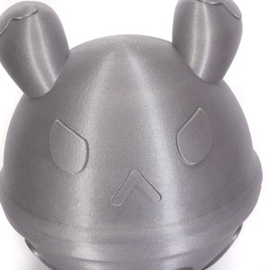 Op de afbeelding: Een grijs 3D-geprint object in de vorm van een hoofd met twee oren en een eenvoudig gezicht. Het object heeft een glad, getextureerd oppervlak.