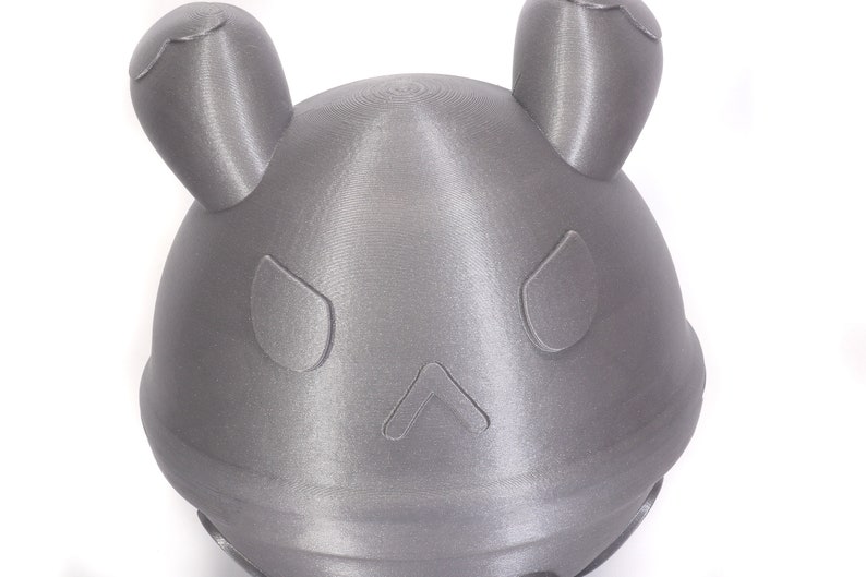 Op de afbeelding: Een grijs 3D-geprint object in de vorm van een hoofd met twee oren en een eenvoudig gezicht. Het object heeft een glad, getextureerd oppervlak.
