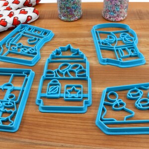 May include: Five blue plastic cookie cutters shaped like different vending machines. The cutters are on a wooden surface.