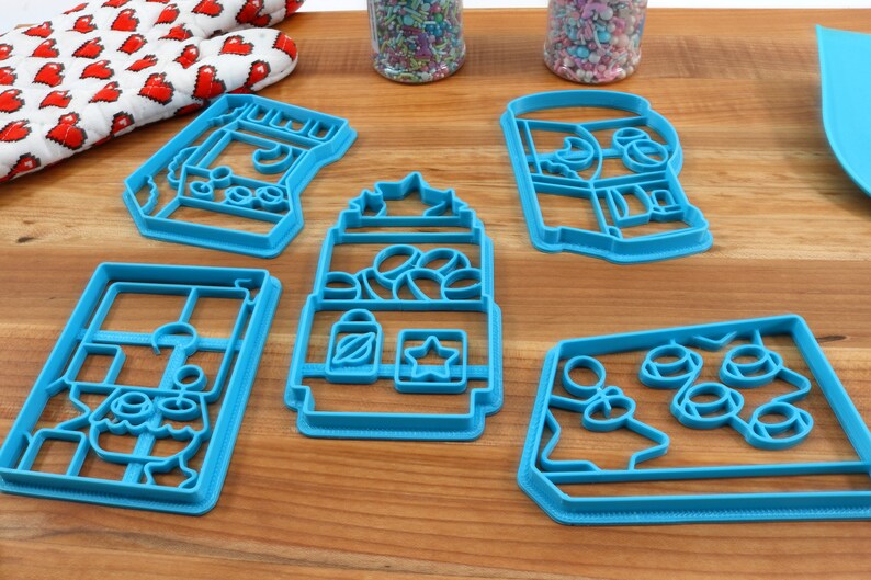May include: Five blue plastic cookie cutters shaped like different vending machines. The cutters are on a wooden surface.