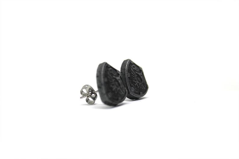 May include: A pair of black metal stud earrings featuring a shield design. The shield has a detailed design and is slightly raised from the earring base.