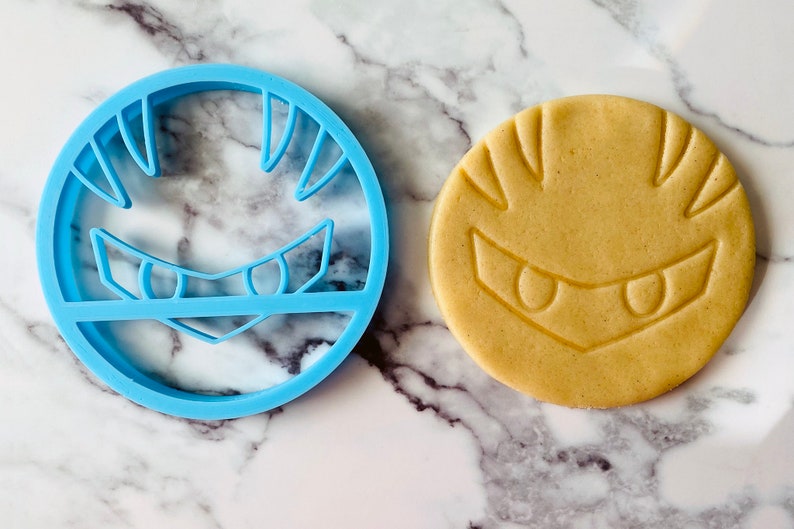 May include: A blue plastic cookie cutter shaped like a video game character's face, next to a light brown cookie with the same design.