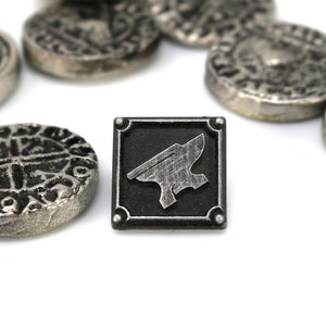 May include: A silver metal token with an anvil design on a black background. The token is surrounded by several other silver metal tokens with intricate designs.