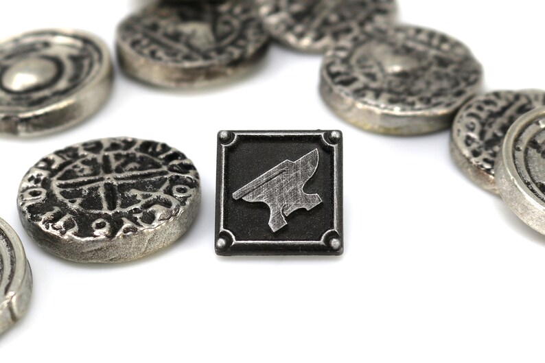 May include: A silver metal token with an anvil design on a black background. The token is surrounded by several other silver metal tokens with intricate designs.