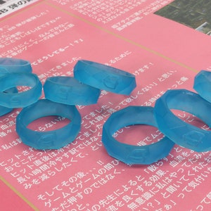 May include: A group of eight translucent blue silicone rings with a textured surface. The rings are arranged in a semi-circle on a pink and white surface.