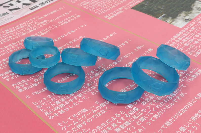 May include: A group of eight translucent blue silicone rings with a textured surface. The rings are arranged in a semi-circle on a pink and white surface.