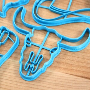 May include: A set of blue plastic cookie cutters in the shape of a cowboy hat, a boot, a star, a bull skull, and a horseshoe.