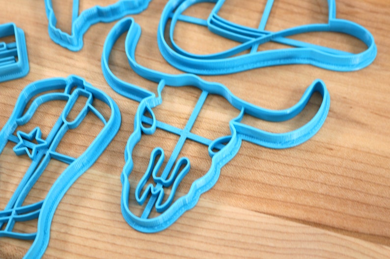 May include: A set of blue plastic cookie cutters in the shape of a cowboy hat, a boot, a star, a bull skull, and a horseshoe.