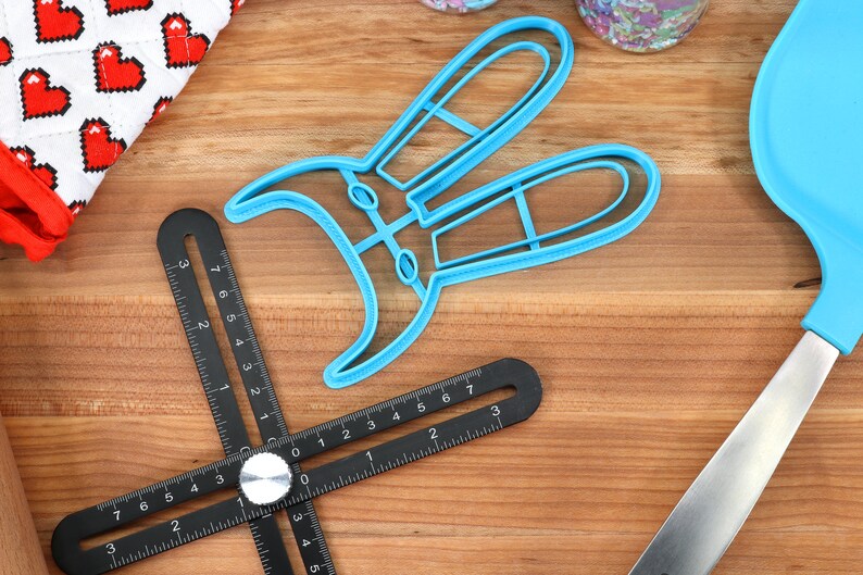 May include: A blue plastic bunny cookie cutter on a wooden surface with a black measuring tool and a blue spatula.