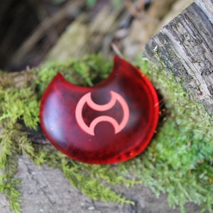 May include: A red crescent-shaped charm with a white symbol in the center. The charm is sitting on a bed of green moss.