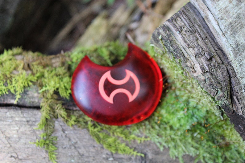 May include: A red crescent-shaped charm with a white symbol in the center. The charm is sitting on a bed of green moss.