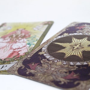 May include: Two tarot cards, one with a woman in a pink and white dress and the other with a large gold star on a dark purple background. The card with the woman has the word "CUP" written on it.