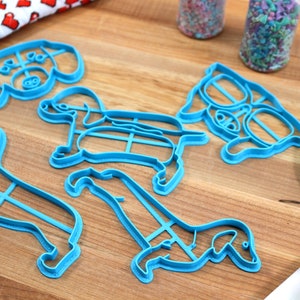 May include: A set of five blue plastic dog-shaped cookie cutters on a wooden surface. The cutters are in various poses, including sitting, standing, and running.
