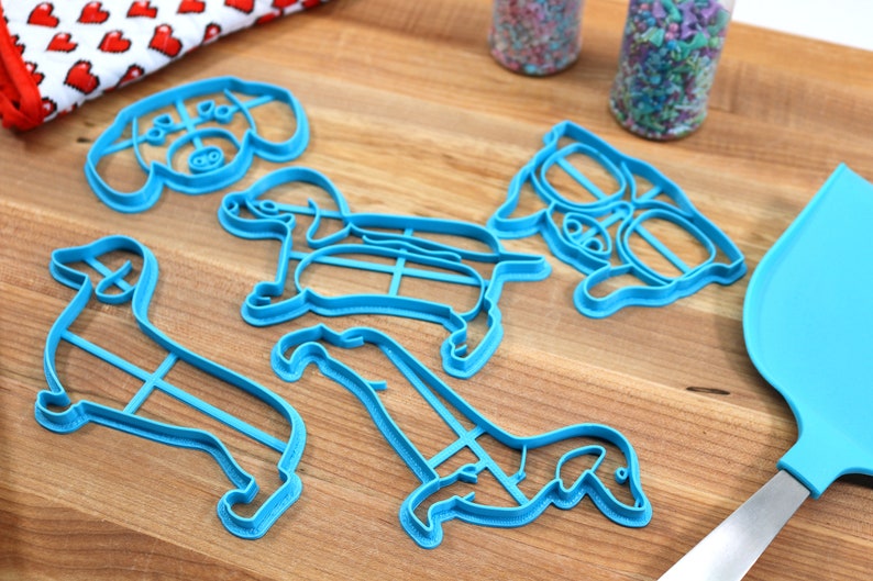 May include: A set of five blue plastic dog-shaped cookie cutters on a wooden surface. The cutters are in various poses, including sitting, standing, and running.