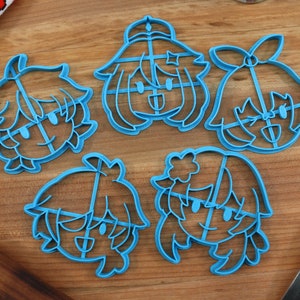 May include: Five blue plastic cookie cutters shaped like cartoon characters with different hairstyles and expressions. The cutters are on a wooden surface.