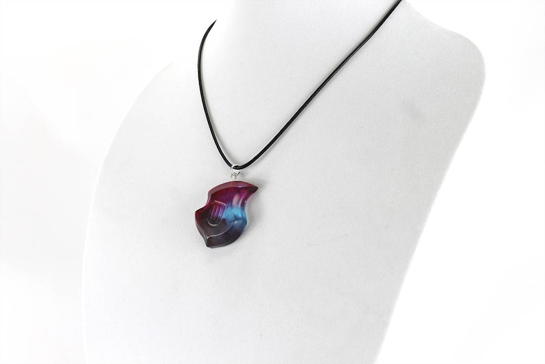 May include: A black cord necklace with a pendant shaped like a lyre. The pendant is made of resin and has a blue, purple, and pink color scheme.