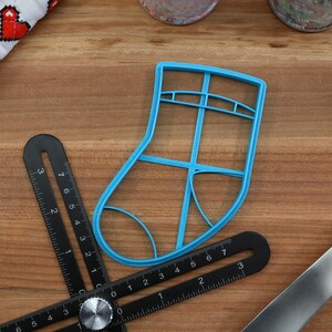 May include: A blue plastic Christmas stocking cookie cutter on a wooden surface with a black measuring tool and a blue spatula.