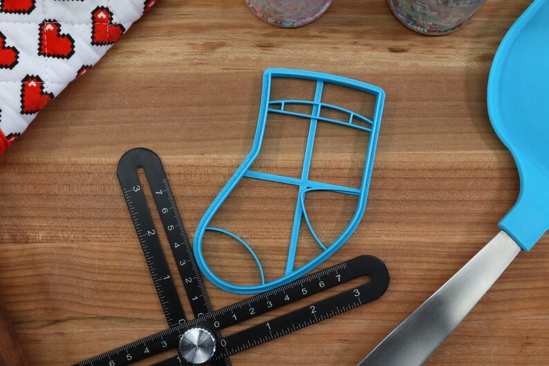 May include: A blue plastic Christmas stocking cookie cutter on a wooden surface with a black measuring tool and a blue spatula.