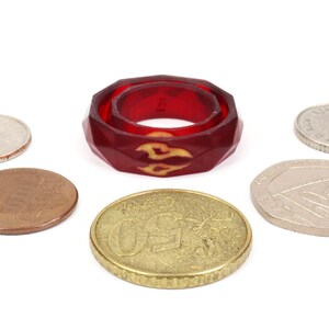 May include: A red, faceted ring with a yellow flame design. The ring is surrounded by six coins.