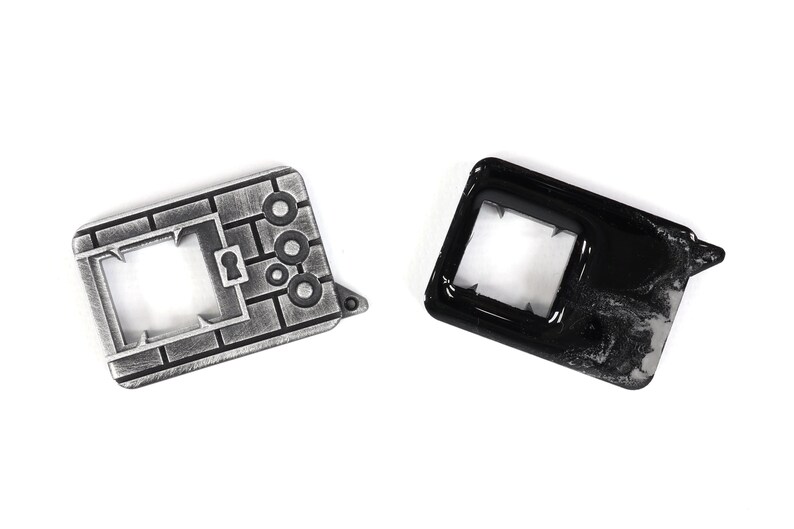 May include: Two metal keychains, one silver and one black. The silver keychain is shaped like a brick wall with a keyhole and three circles. The black keychain is a simple rectangle with a square cutout.