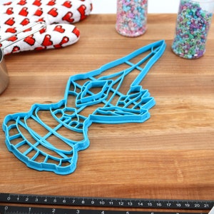 May include: A blue plastic cookie cutter shaped like a stylized bird with a long, pointed beak. The cutter is on a wooden surface with a ruler, measuring cup, and other baking supplies.