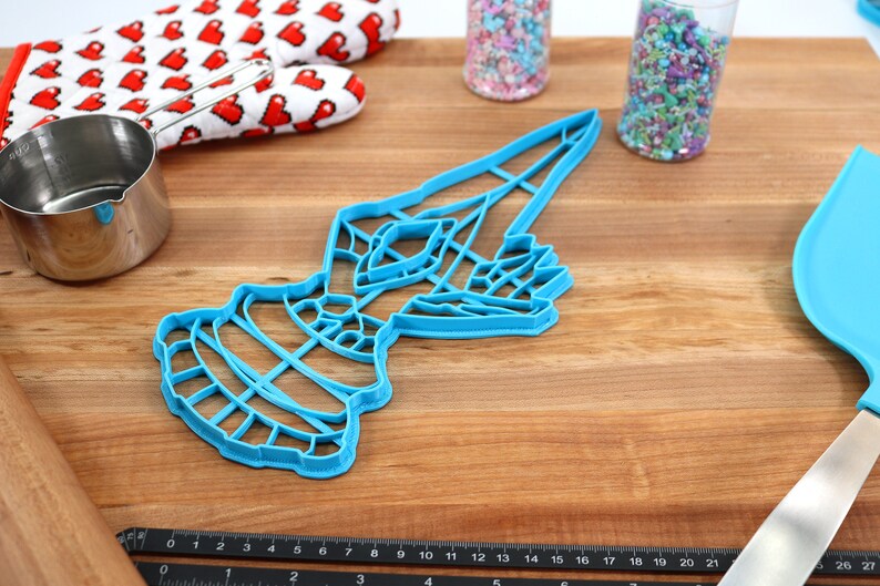 May include: A blue plastic cookie cutter shaped like a stylized bird with a long, pointed beak. The cutter is on a wooden surface with a ruler, measuring cup, and other baking supplies.
