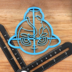 May include: A blue plastic cookie cutter shaped like a cartoon character with large eyes and a smile. The cutter is on a wooden surface with a black ruler.