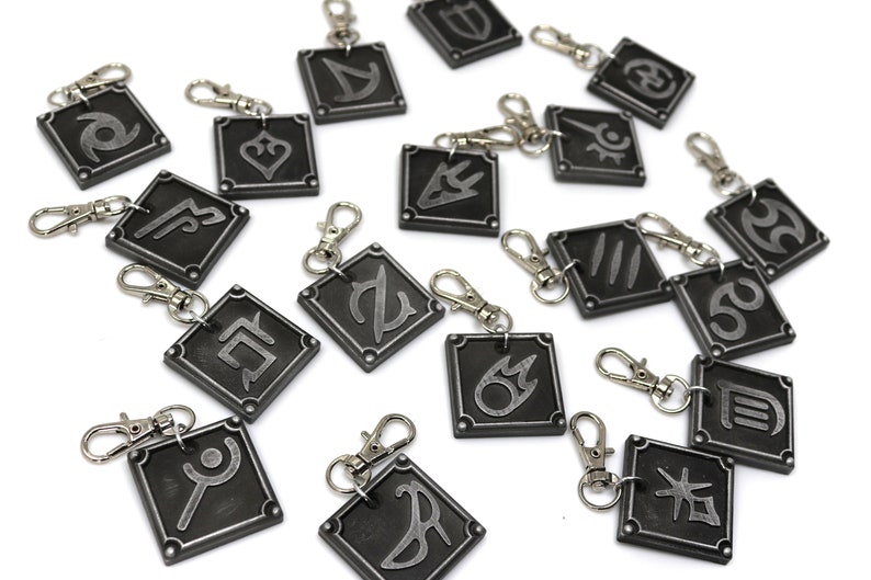 May include: Set of 16 silver and black metal keychains with different symbols. Each keychain features a square-shaped charm with a silver symbol on a black background.
