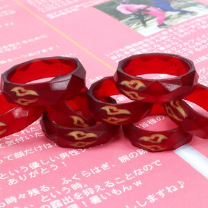 May include: A stack of six red resin rings with a gold design. The rings are stacked on a pink surface.