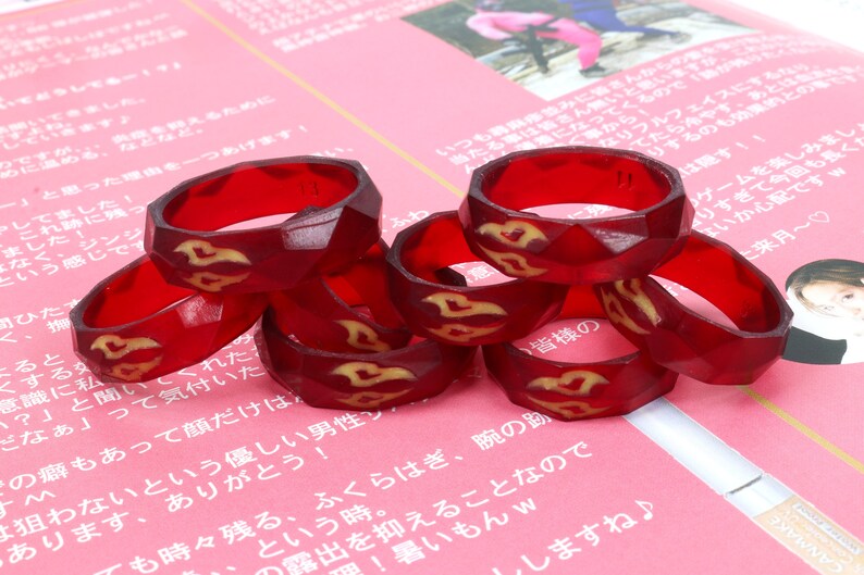 May include: A stack of six red resin rings with a gold design. The rings are stacked on a pink surface.