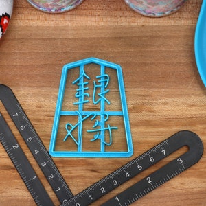 May include: A light blue plastic cookie cutter with Chinese characters is centered on a wooden surface. Black rulers and a blue spatula are also visible. A white oven mitt with red pixelated hearts is in the upper left corner.