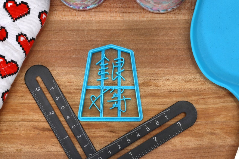 May include: A light blue plastic cookie cutter with Chinese characters is centered on a wooden surface. Black rulers and a blue spatula are also visible. A white oven mitt with red pixelated hearts is in the upper left corner.